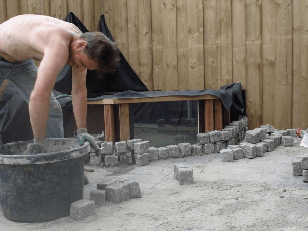 Aanleg keermuur van klinkers rond vijver met glazen wand.