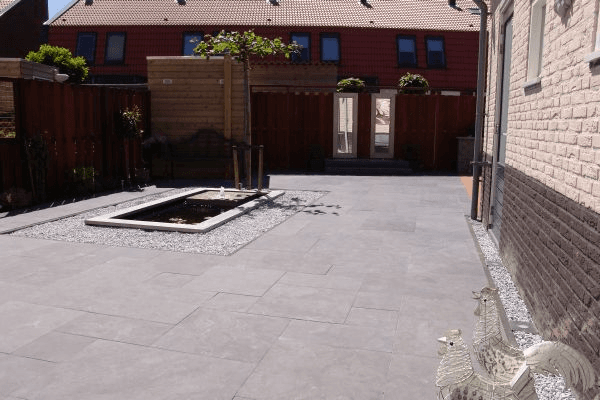 Achtertuin met grijze tegels, rechthoekige vijver met fontein en wit grind.