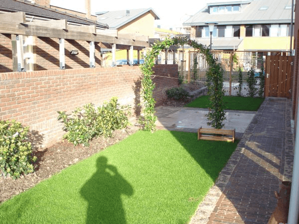 Smalle achtertuin met gazon, bakstenen muur, houten pergola, boog met klimplant en terras met grote tegels.
