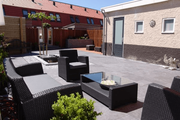 Achtertuinterras met donkergrijze tegels, loungeset, rechthoekige vijver met fontein, bijgebouw en houten schutting.