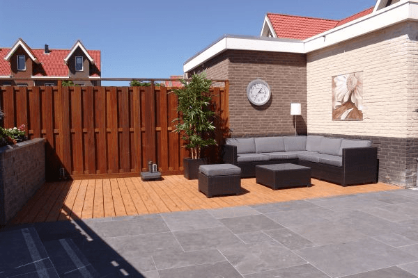 Loungehoek op houten vlonder naast donkere bestrating; bakstenen gevel met klok en schilderij, houten schutting.