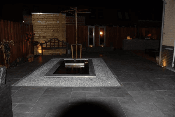 Tuin ’s avonds: centrale waterpartij in grindbed, donkere tegels, sfeerverlichting en houten bank tegen schutting.