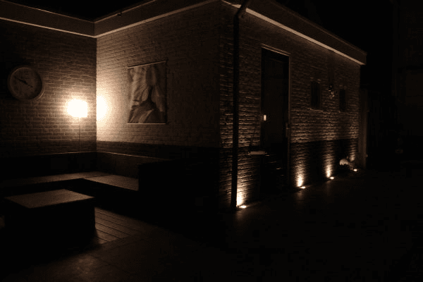 Avondbeeld terras bij twee-kleuren bakstenen muur, loungeset, wandlamp en grondspots tegen de gevel.